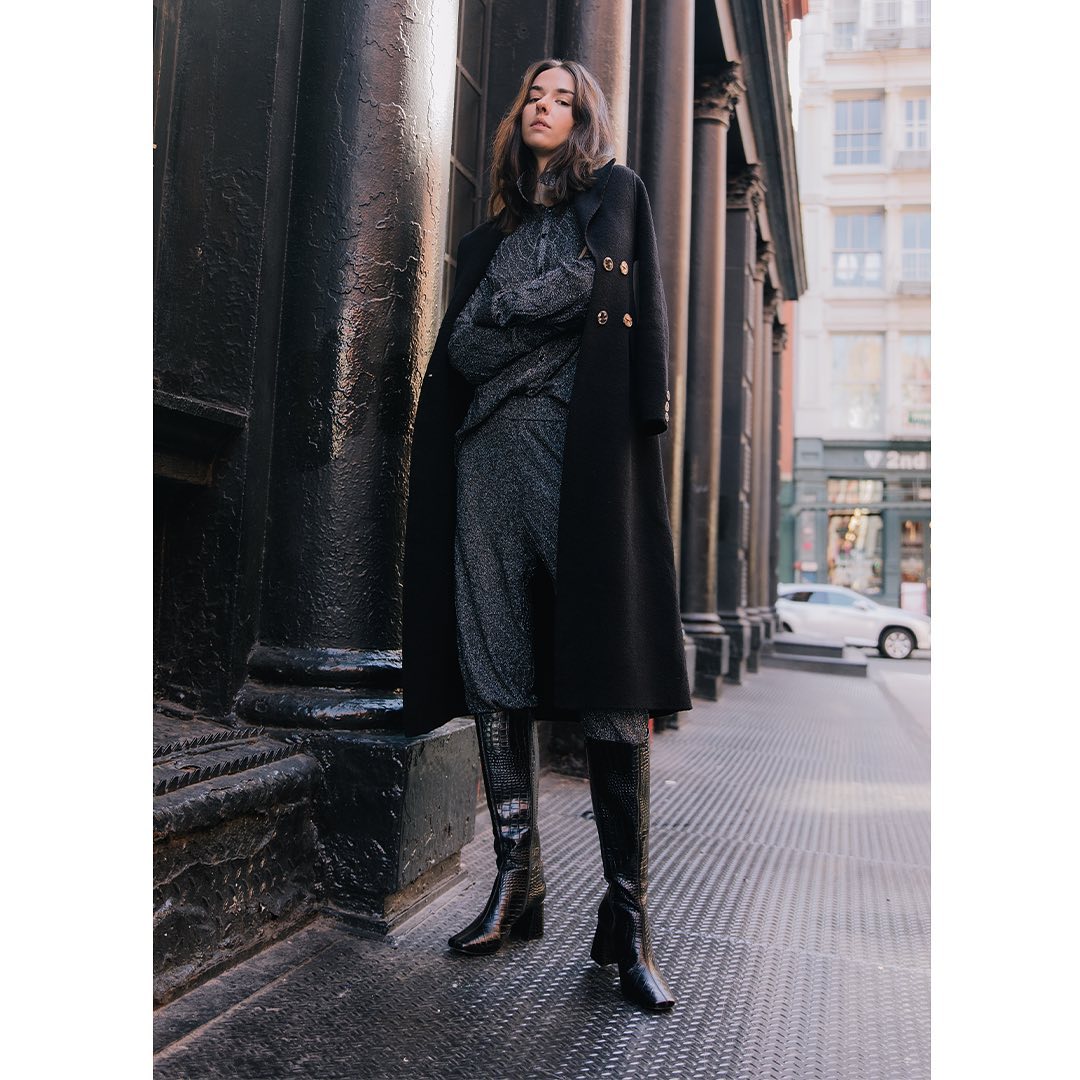 Woman in a black coat and pants standing on a city street.