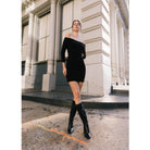 Woman in a black off-shoulder dress and knee-high boots standing on a city street.